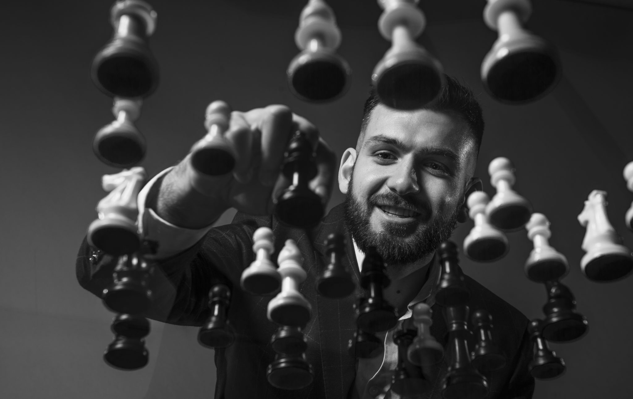 A man in a suit smiling as he reaches through a suspended chessboard, adjusting a piece. The black and white image conveys focus and strategy.