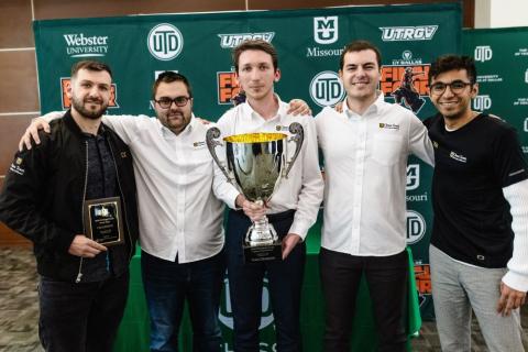 A group of five men poses with a large trophy in front of a green backdrop featuring university logos. One man holds a plaque. All are smiling.